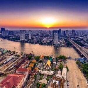 Aerial view of Bangkok city skyline with Chao Phraya River at sunset, featuring skyscrapers, bridges, and colorful temples.