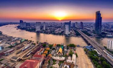 Aerial view of Bangkok city skyline with Chao Phraya River at sunset, featuring skyscrapers, bridges, and colorful temples.