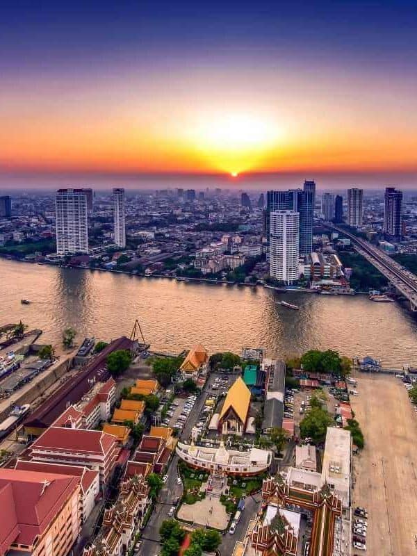 Aerial view of Bangkok city skyline with Chao Phraya River at sunset, featuring skyscrapers, bridges, and colorful temples.
