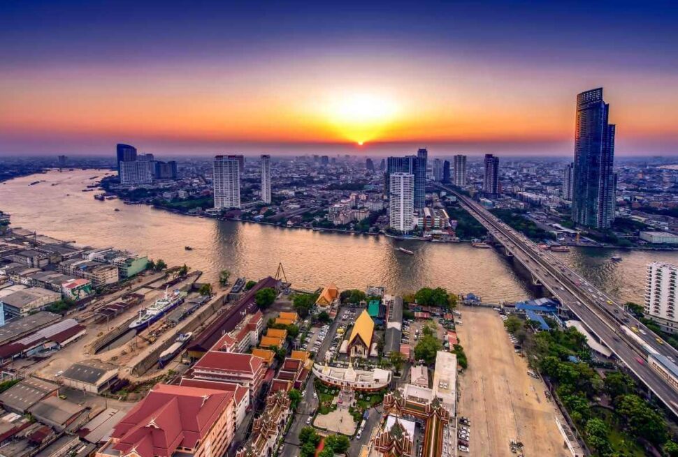 Aerial view of Bangkok city skyline with Chao Phraya River at sunset, featuring skyscrapers, bridges, and colorful temples.