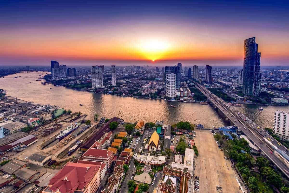 Aerial view of Bangkok city skyline with Chao Phraya River at sunset, featuring skyscrapers, bridges, and colorful temples.