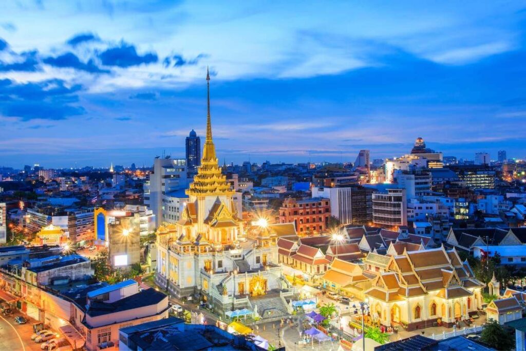 Wat Traimit Golden Buddha Temple illuminated at night in Bangkok, Thailand, with city lights and modern skyline in the background.