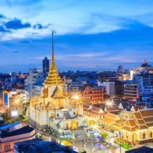 Wat Traimit Golden Buddha Temple illuminated at night in Bangkok, Thailand, with city lights and modern skyline in the background.