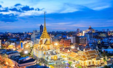 Wat Traimit Golden Buddha Temple illuminated at night in Bangkok, Thailand, with city lights and modern skyline in the background.