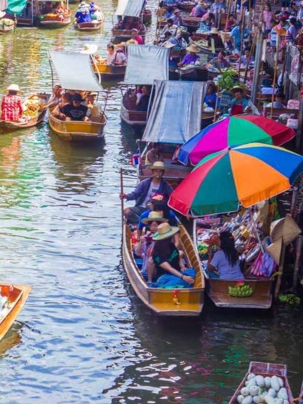 Colorful floating market in Thailand with vendors selling goods from wooden boats under umbrellas on a busy canal.