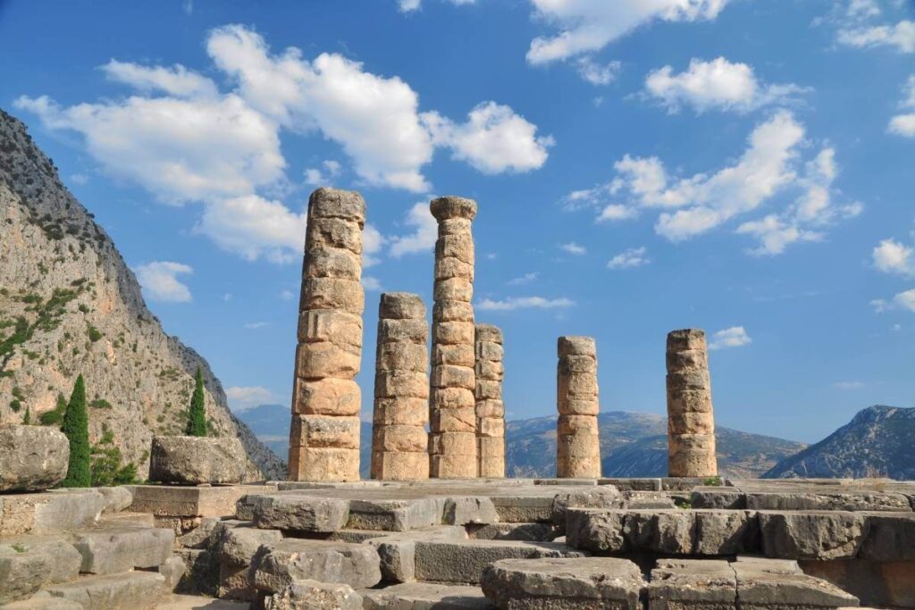 Stone columns of the Temple of Apollo at Delphi, Greece, set against mountains and a bright blue sky.