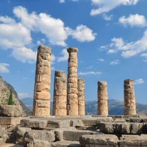 Stone columns of the Temple of Apollo at Delphi, Greece, set against mountains and a bright blue sky.