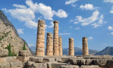 Stone columns of the Temple of Apollo at Delphi, Greece, set against mountains and a bright blue sky.