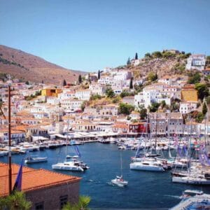 Scenic view of Hydra Island’s main port in Greece with sailboats, colorful houses, and hillside landscape under a clear sky.