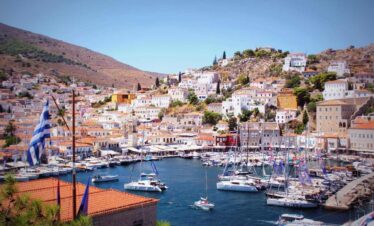Scenic view of Hydra Island’s main port in Greece with sailboats, colorful houses, and hillside landscape under a clear sky.