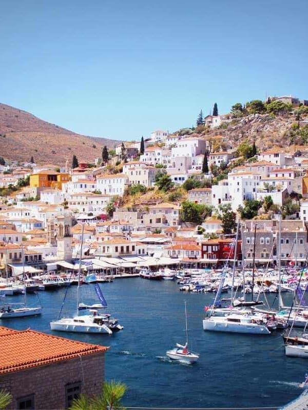 Vista panorámica del puerto principal de la isla de Hydra en Grecia, con veleros, casas coloridas y paisaje de laderas bajo un cielo despejado.