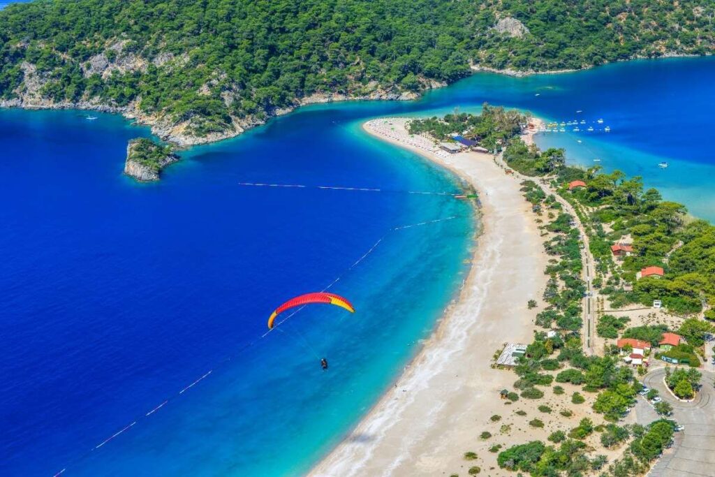 Aerial view of Ölüdeniz Beach in Turkey with turquoise water, lush green hills, and a paraglider soaring above the blue lagoon.