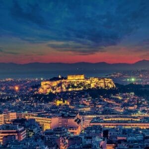 Illuminated Acropolis of Athens at dusk with city lights and colorful sunset sky in the background.