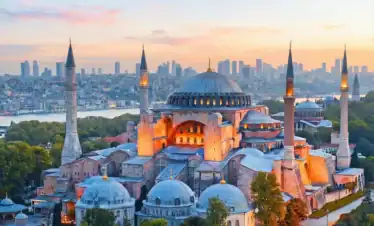 Panoramic sunset view of the Hagia Sophia Grand Mosque in Istanbul, showcasing its iconic massive dome and minarets against a vibrant orange sky, with the Bosphorus strait visible in the background.