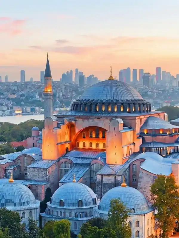 Panoramic sunset view of the Hagia Sophia Grand Mosque in Istanbul, showcasing its iconic massive dome and minarets against a vibrant orange sky, with the Bosphorus strait visible in the background.