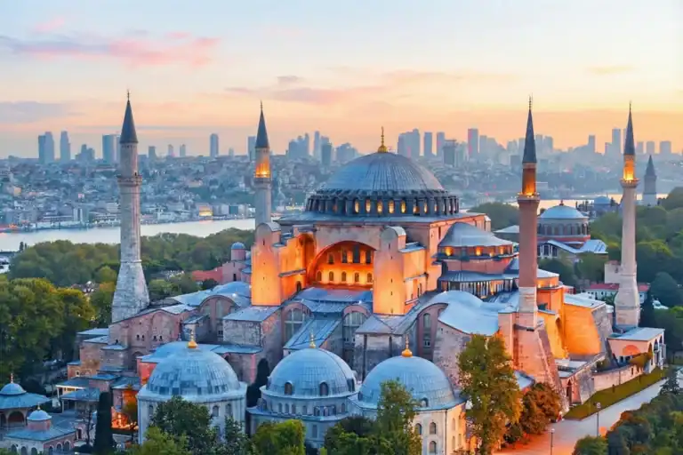 Panoramic sunset view of the Hagia Sophia Grand Mosque in Istanbul, showcasing its iconic massive dome and minarets against a vibrant orange sky, with the Bosphorus strait visible in the background.