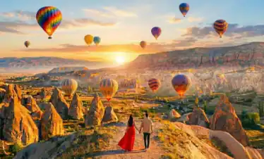 A couple holding hands walks along a panoramic trail at sunrise, looking out over the unique rock formations of Cappadocia, Turkey, as dozens of colorful hot air balloons float in the golden sky.