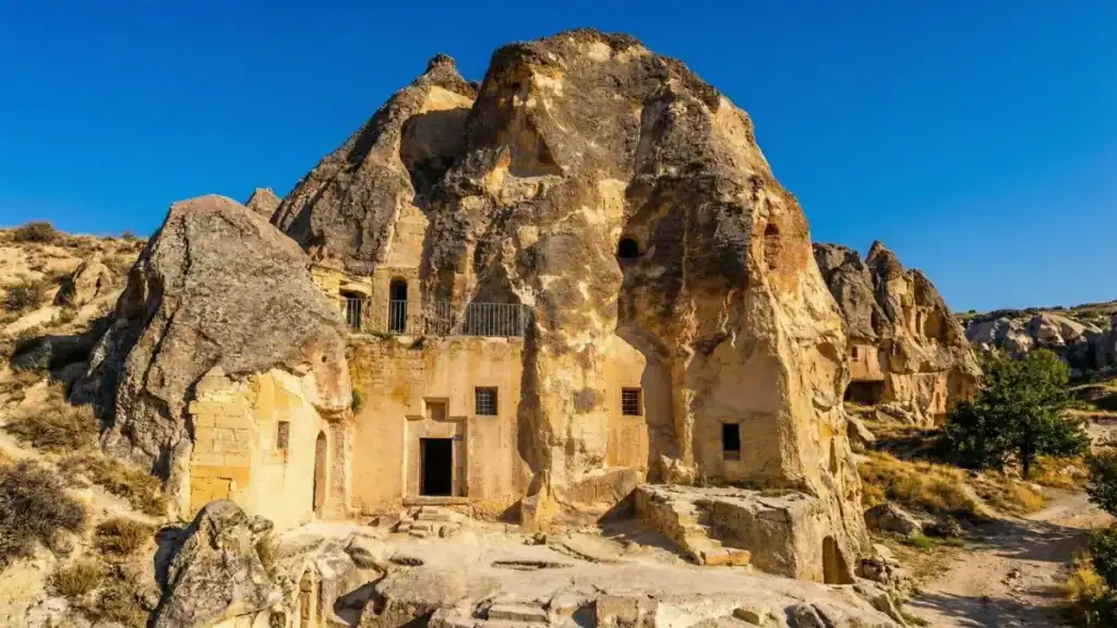 A wide-angle landscape photograph of the ancient rock-cut Keslik Monastery complex in Cappadocia, Turkey, showing carved entrances, windows, stone steps, and a metal railing under a clear deep blue sky.
