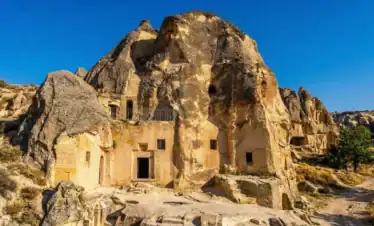 A wide-angle landscape photograph of the ancient rock-cut Keslik Monastery complex in Cappadocia, Turkey, showing carved entrances, windows, stone steps, and a metal railing under a clear deep blue sky.