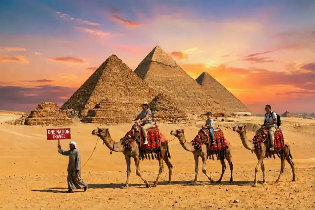 One Nation Travel guide holding a red sign leading an Egypt tour with tourists riding camels in front of the Giza Pyramids at sunset