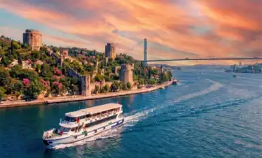 A breathtaking sunset view of the Rumeli Fortress and Bosphorus Bridge in Istanbul, with a tour boat cruising on the vivid orange and blue waters.