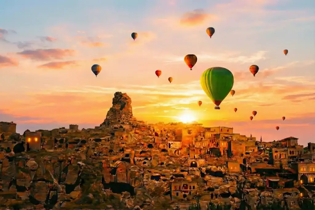 After 500 Cappadocia Sunrise Tours, I Finally Know Why 80% Fail 4 A wide shot of the rock-hewn town and Ortahisar Castle in Cappadocia at sunrise, with numerous hot air balloons, including a prominent green one, floating in the golden-hour sky.