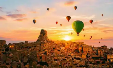 A wide shot of the rock-hewn town and Ortahisar Castle in Cappadocia at sunrise, with numerous hot air balloons, including a prominent green one, floating in the golden-hour sky.