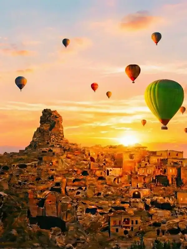 A wide shot of the rock-hewn town and Ortahisar Castle in Cappadocia at sunrise, with numerous hot air balloons, including a prominent green one, floating in the golden-hour sky.