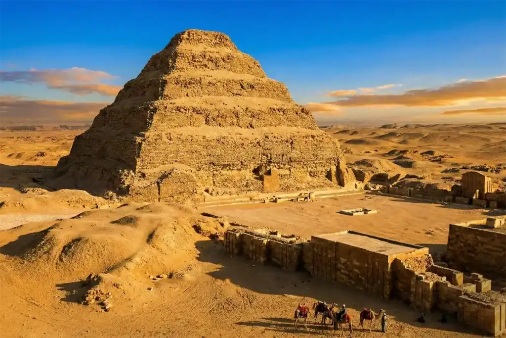 The Step Pyramid of Djoser rising from the desert sands of Saqqara, Egypt, under a blue and orange sunset sky. In the fore...