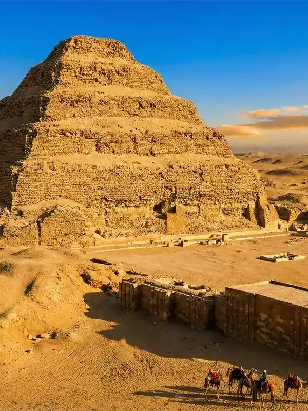 The Step Pyramid of Djoser rising from the desert sands of Saqqara, Egypt, under a blue and orange sunset sky. In the fore...