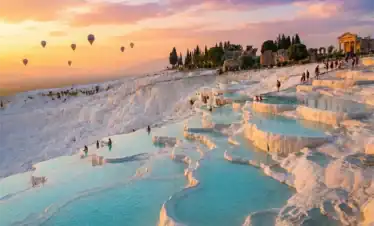Sunset view of Pamukkale white travertine terraces and turquoise thermal pools in Turkey with hot air balloons floating above.