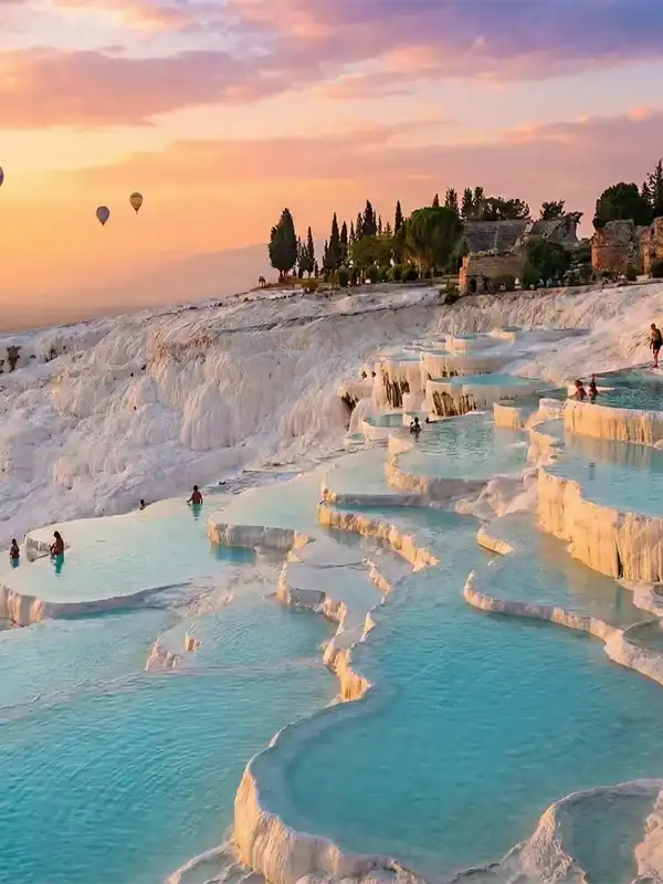 Sunset view of Pamukkale white travertine terraces and turquoise thermal pools in Turkey with hot air balloons floating above.