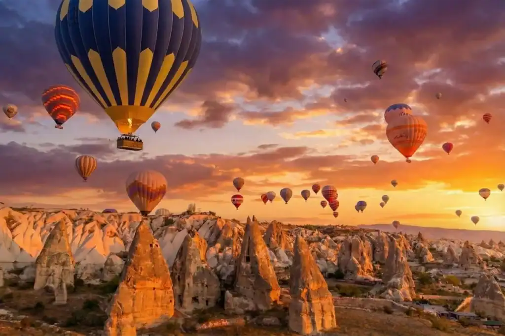 A prominent blue and yellow striped hot air balloon flying alongside dozens of others during sunrise over the unique fairy chimney rock formations in Cappadocia, Turkey, a key highlight of the 2-Day Cappadocia Tour from Istanbul.