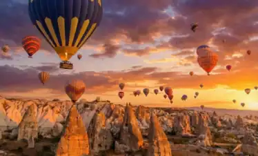 A prominent blue and yellow striped hot air balloon flying alongside dozens of others during sunrise over the unique fairy chimney rock formations in Cappadocia, Turkey, a key highlight of the 2-Day Cappadocia Tour from Istanbul.