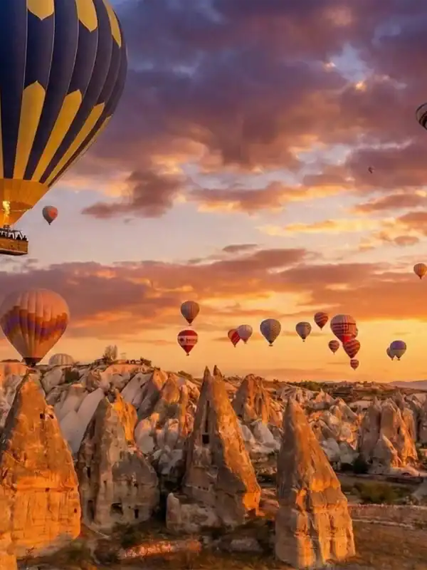A prominent blue and yellow striped hot air balloon flying alongside dozens of others during sunrise over the unique fairy chimney rock formations in Cappadocia, Turkey, a key highlight of the 2-Day Cappadocia Tour from Istanbul.