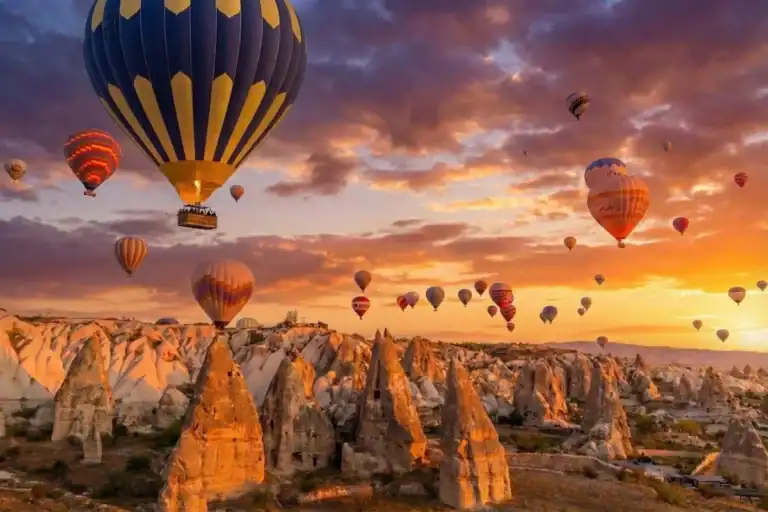 Istanbul to Cappadocia: How to Plan the Perfect Trip 1 A prominent blue and yellow striped hot air balloon flying alongside dozens of others during sunrise over the unique fairy chimney rock formations in Cappadocia, Turkey, a key highlight of the 2-Day Cappadocia Tour from Istanbul.