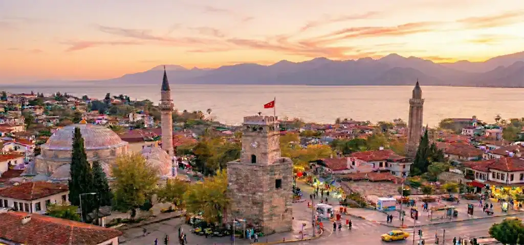Vista panorámica de la puesta de sol sobre el casco antiguo de Antalya Kaleici, con su histórica torre del reloj, minaretes y un barco turístico en el mar Mediterráneo.