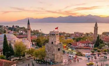 Panoramic sunset view of Antalya Kaleici old town featuring the historic clock tower, minarets, and a sightseeing boat on the Mediterranean Sea.