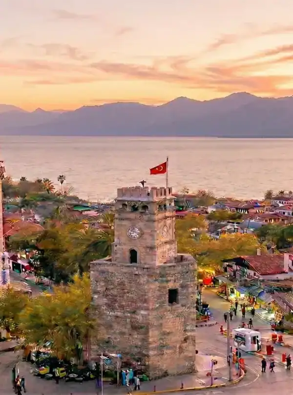Panoramic sunset view of Antalya Kaleici old town featuring the historic clock tower, minarets, and a sightseeing boat on the Mediterranean Sea.