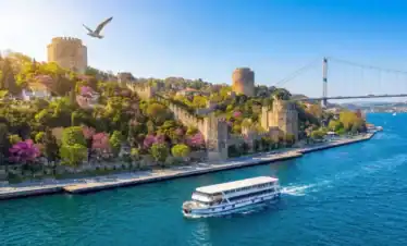 Scenic Bosphorus cruise boat sailing past the historical Rumeli Fortress and bridge in Istanbul on a sunny day.