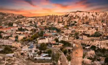 Panoramic sunrise view from Esentepe viewpoint overlooking Goreme village in Cappadocia, featuring tourists on a lookout deck, hot air balloons in the distance, and dramatic orange morning skies.