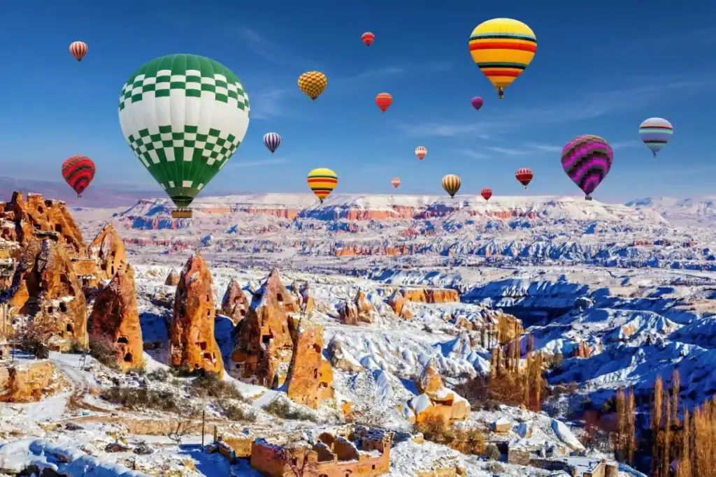 Colorful hot air balloons float above snow-covered fairy chimneys and ancient cave dwellings in wintery Cappadocia, Turkey, set against a bright blue sky.