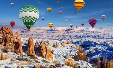 Colorful hot air balloons float above snow-covered fairy chimneys and ancient cave dwellings in wintery Cappadocia, Turkey, set against a bright blue sky.