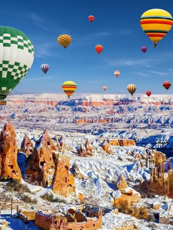 Colorful hot air balloons float above snow-covered fairy chimneys and ancient cave dwellings in wintery Cappadocia, Turkey, set against a bright blue sky.