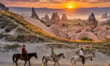 Three riders in red, green, and white clothing on horseback, exploring the majestic fairy chimneys and valleys of Cappadocia during a vibrant golden sunset.