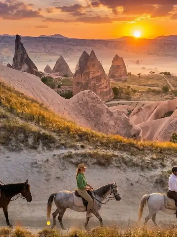 Three riders in red, green, and white clothing on horseback, exploring the majestic fairy chimneys and valleys of Cappadocia during a vibrant golden sunset.