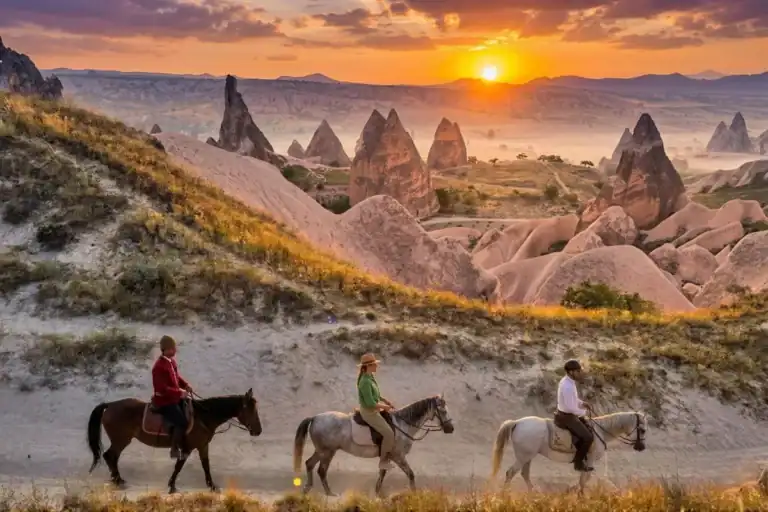Three riders in red, green, and white clothing on horseback, exploring the majestic fairy chimneys and valleys of Cappadocia during a vibrant golden sunset.