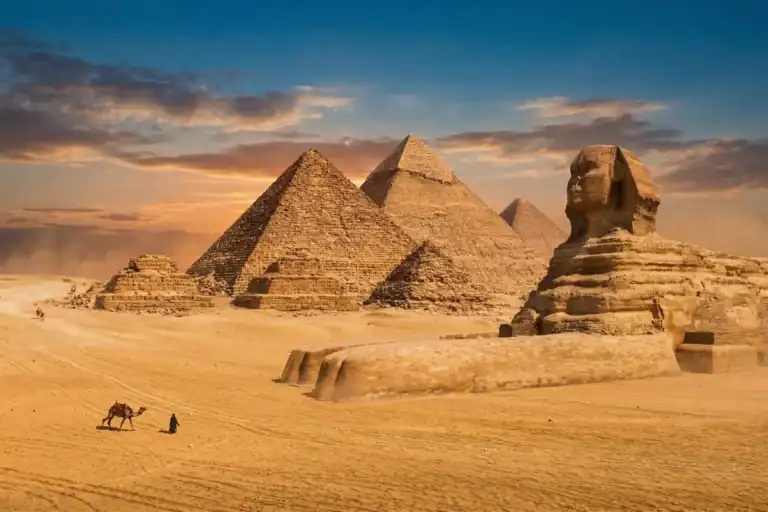 Breathtaking view of the Great Sphinx and Pyramids of Giza in Egypt. A local bedouin walks a camel across the golden desert sand under a clear blue sky. A must-see historic wonder for travelers.