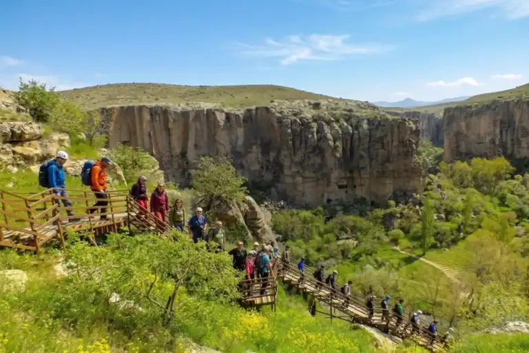 Pemandangan panorama sekumpulan pendaki menuruni tangga kayu yang berliku ke dalam gaung Lembah Ihlara di Cappadocia, Turki. Permukaan tebing yang bertentangan dipenuhi dengan kediaman gua purba yang dipahat dari batu dan gereja, di bawah langit biru yang sebahagiannya mendung. Dasar lembah di bawahnya subur dan menghijau dengan pokok-pokok.