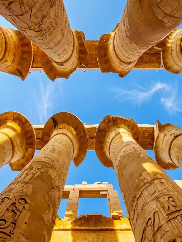 Imposing view looking up at the hieroglyph-carved stone columns of the Great Hypostyle Hall in Karnak Temple, Luxor. Clear blue sky visible between the massive columns.